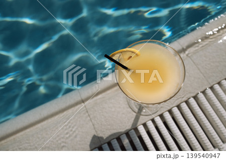 An elegant glass with a margarita cocktail with a slice of lemon and a straw on the side of the pool. Top view. 139540947