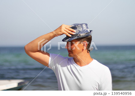 Portrait of middle-aged male tourist of Brazilian appearance in white T-shirt and summer hat against backdrop of sea, copy space Portrait of middle-aged male tourist of Brazilian appearance in white T-shirt and summer hat against backdrop of sea, copy space 139540956