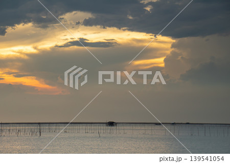 Warm light pierces dense clouds over a quiet shoreline with fish farm stakes and a small hut, creating a soft horizon and reflective water under an approaching storm. 139541054