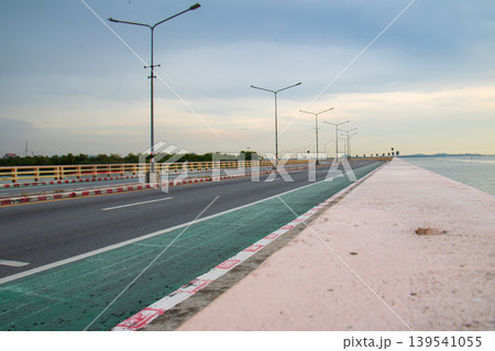 Long perspective of a quiet seaside roadway with evenly spaced streetlights, painted lane markings, and calm water beside the embankment under overcast evening light. 139541055