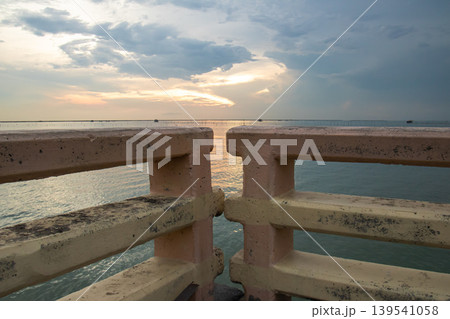 Concrete guardrail frames a tranquil ocean view as soft sunset light reflects on the water, creating a peaceful coastal atmosphere ideal for travel, relaxation, and outdoor concepts. Concrete guardrail frames a tranquil ocean view as soft sunset light reflects on the water, creating a peaceful coastal atmosphere ideal for travel, relaxation, and outdoor concepts. 139541058