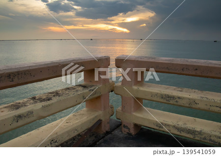 Aged concrete railing overlooks a calm sea under a glowing sunset sky, with soft reflections on the water and layered clouds creating a serene coastal atmosphere. 139541059