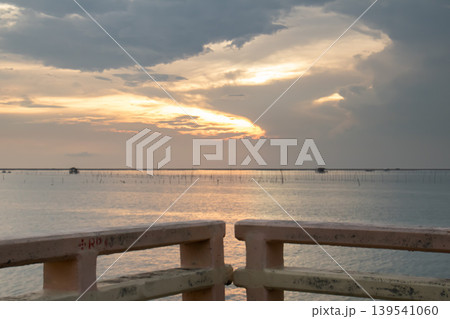 Soft sunset light reflects across a quiet sea dotted with fishing poles, viewed from a weathered concrete railing, creating a tranquil coastal scene with warm tones and gentle atmosphere. 139541060