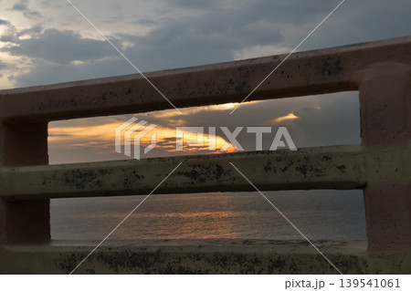Moody sunset framed by weathered metal railings, with warm golden light reflecting on a calm sea under dramatic clouds. Concept of solitude, barrier, travel, and coastal atmosphere. 139541061