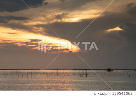 Warm sunset light spreads across shallow coastal waters dotted with fishing stakes and a distant hut. Dramatic clouds create a serene yet powerful seascape, evoking calm, livelihood, and coastal life. 139541062