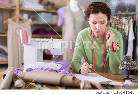 Sad female dressmaker sitting over blank sheet of paper in sewing studio Sad female dressmaker sitting over blank sheet of paper in sewing studio 139541646