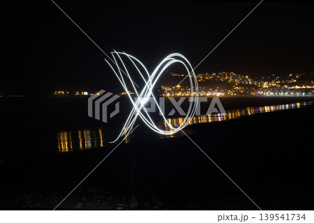 Abstract light painting trail on dark beach at night with city lights background Abstract light painting trail on dark beach at night with city lights background 139541734