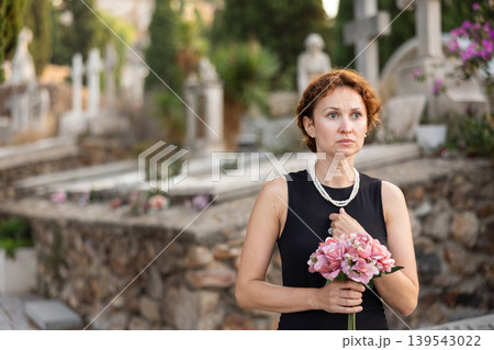 Woman in mourning black dress standing in cemetery with flowers 139543022