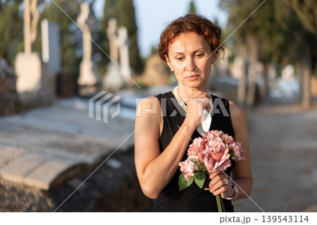 Sad woman in black dress with bouquet of flowers and handkerchief in hands, stands in cemetery 139543114