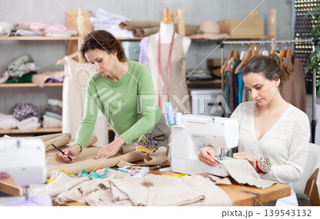 Female tailor sews on a sewing machine while another tailor draws outline of a future dress on paper Female tailor sews on a sewing machine while another tailor draws outline of a future dress on paper 139543132