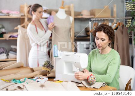Female dressmaker sewing at machine while colleague working with mannequin Female dressmaker sewing at machine while colleague working with mannequin 139543140