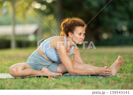 Woman practicing hatha yoga in garden, stretching in an asymmetrical seated asana Parivritta Janu Sirsasana 139543141