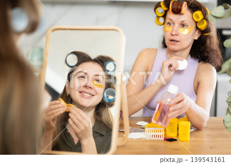 Woman in colorful curlers applying under eye patches by mirror with bestie 139543161