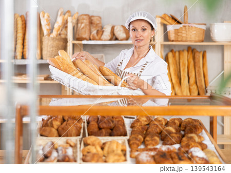 Positive female bakery employee offering bread and different baguettes for sale Positive female bakery employee offering bread and different baguettes for sale 139543164