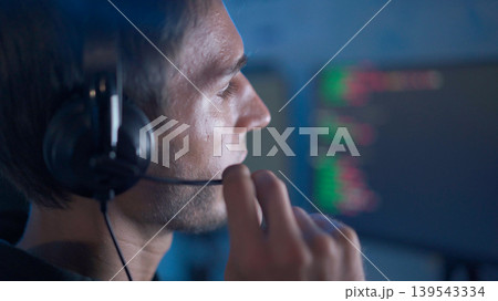 Software developer analyzing code on computer monitors while wearing headset in dark workspace 139543334