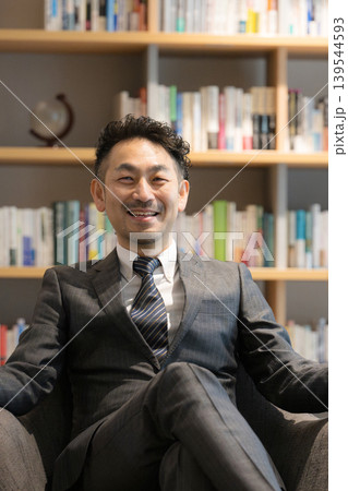 社長室のような本棚の前で座る、笑顔でカメラ目線の経営者ポートレート成功と信頼の仕事イメージ 139544593