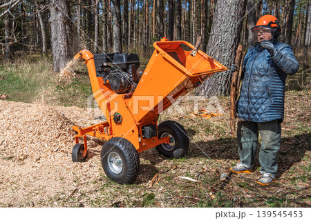 Wood chipper at work in forest, woman in safety gear operating machine, wood chips flying Wood chipper at work in forest, woman in safety gear operating machine, wood chips flying 139545453