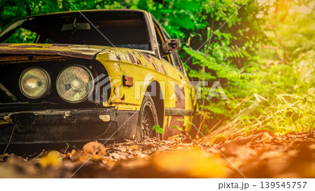Old wrecked car in vintage style. Abandoned rusty yellow car in the forest. Closeup front view headlights of rusty wrecked abandoned car on blurred green tree background . Art of abandoned used car. 139545757