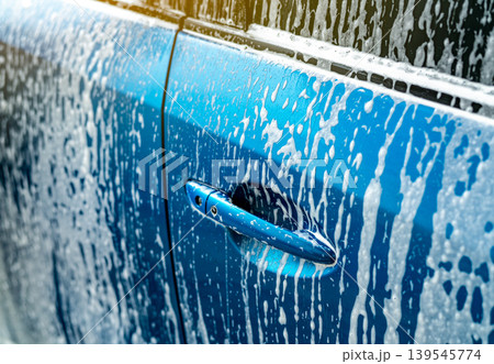 Blue car covered with white soap foam during professional carwash service in auto care business. Vehicle cleaning process before waxing for glossy shine and maintenance quality in modern garage. Blue car covered with white soap foam during professional carwash service in auto care business. Vehicle cleaning process before waxing for glossy shine and maintenance quality in modern garage. 139545774