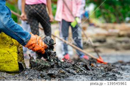 Adult and children volunteers collecting garbage on the sea beach. Beach environment pollution. Tidying up rubbish on beach. People wear orange gloves picking garbage up in to yellow bag. 139545961