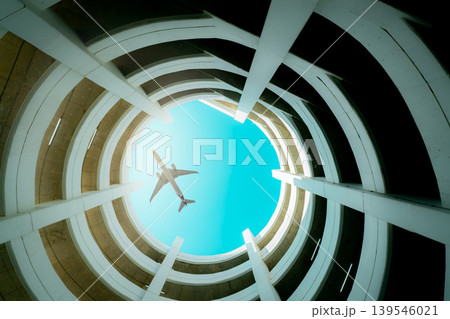 Bottom view multi-story car park building with plane flying above building. Air logistic. Aviation and travel business.  Architecture of spiral curve building. Airline business. Tourism transportation 139546021