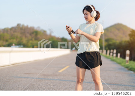 Asian woman with headphones using smartphone in the park. Female runner adjusting music or tracking app before jogging. Technology, mobile device and fitness workout concept in morning 139546202