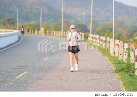 Active Asian woman jogging with a towel around her neck in the park at morning. Female runner exercising in summer for healthy lifestyle and fitness workout outdoors. Wellness and sport concepts Active Asian woman jogging with a towel around her neck in the park at morning. Female runner exercising in summer for healthy lifestyle and fitness workout outdoors. Wellness and sport concepts 139546216