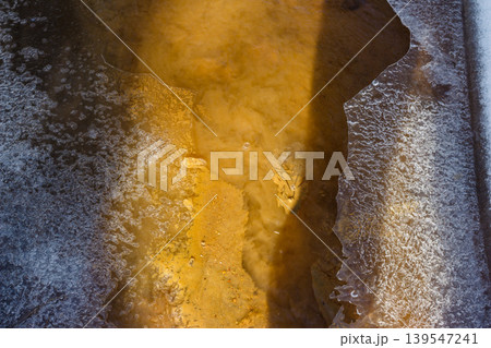 Murky brown stream water reveals dirty silt and exposed creek bed, bordered by jagged, melting ice. A raw, textural scene of early spring thaw 139547241
