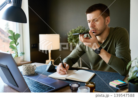 Man recording voice message on smartphone and writing notes at home desk Man recording voice message on smartphone and writing notes at home desk 139547588