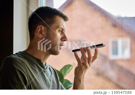 Man using voice assistant on smartphone by window at home 139547589