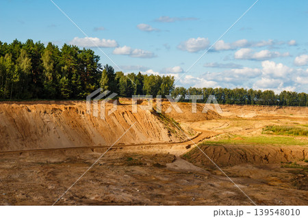Vast sand quarry with steep, eroded slopes and sandy terrain. Dense forest borders the pit under a bright sky, showcasing an open-cast landscape Vast sand quarry with steep, eroded slopes and sandy terrain. Dense forest borders the pit under a bright sky, showcasing an open-cast landscape 139548010