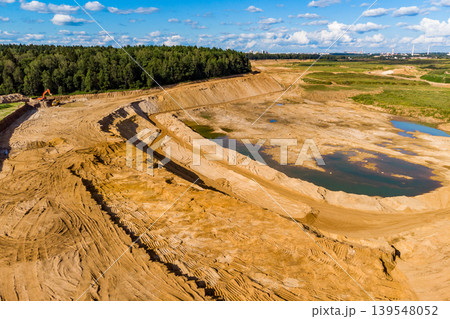 Expansive sand excavation site with vast sandy terrain, deep pits, and water pools. Nearby forest, distant urban skyline, and a sunny, cloudy sky Expansive sand excavation site with vast sandy terrain, deep pits, and water pools. Nearby forest, distant urban skyline, and a sunny, cloudy sky 139548052