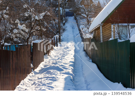 Winding snowy rural path, deeply trodden, ascends between rustic fences and homes on a bright winter day 139548536