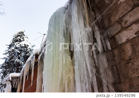 Dramatic, thick icicles cascade down the frosty brick and concrete walls of an old building, capturing winter's stark, frozen beauty. Clear sky and snowy tree Dramatic, thick icicles cascade down the frosty brick and concrete walls of an old building, capturing winter's stark, frozen beauty. Clear sky and snowy tree 139549296