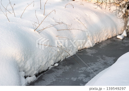 A tranquil frozen stream flows between pristine snow-covered banks. Delicate winter branches pierce the fresh white blanket, capturing the serene essence of a frosty day A tranquil frozen stream flows between pristine snow-covered banks. Delicate winter branches pierce the fresh white blanket, capturing the serene essence of a frosty day 139549297