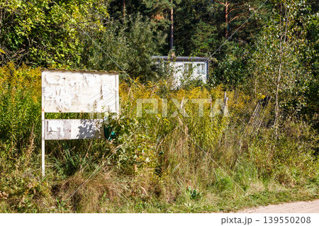 Weathered bulletin board in an overgrown rural setting, covered with ripped papers. A small white house peeks through lush greenery and tall wild grasses under a clear sky Weathered bulletin board in an overgrown rural setting, covered with ripped papers. A small white house peeks through lush greenery and tall wild grasses under a clear sky 139550208