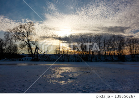 Stark silhouettes of bare trees line a frozen, snow-dusted river at sunset. The tranquil winter scene showcases nature's raw, cold beauty under a warm, glowing sky 139550267
