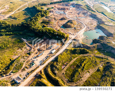 Aerial view of an active open-pit quarry with multiple excavated pits and turquoise water pools. A dirt road snakes through the industrial landscape, showing ongoing resource extraction and Aerial view of an active open-pit quarry with multiple excavated pits and turquoise water pools. A dirt road snakes through the industrial landscape, showing ongoing resource extraction and 139550271