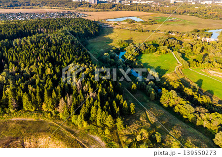 Aerial view of a sprawling green forest bordering a meandering river, with fields and distant urban sprawl under warm sunlight. A serene natural landscape in late summer 139550273
