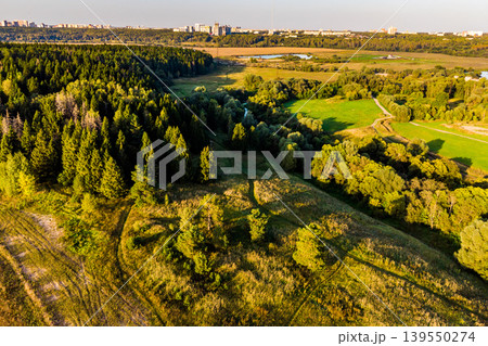 Lush forest and rolling hills bathed in golden hour glow, with winding paths leading through verdant foliage. A distant city skyline hints at urban proximity to this peaceful nature haven 139550274