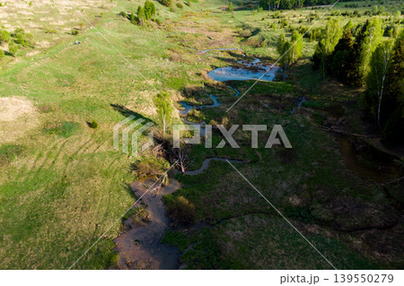 Meandering stream flows through a lush green valley, creating small ponds and wetlands. Bright sun illuminates fresh spring foliage and distant hills Meandering stream flows through a lush green valley, creating small ponds and wetlands. Bright sun illuminates fresh spring foliage and distant hills 139550279