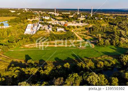 Drone shot of a bustling industrial complex with towering stacks, surrounded by verdant fields and a meandering river, bathed in warm golden light Drone shot of a bustling industrial complex with towering stacks, surrounded by verdant fields and a meandering river, bathed in warm golden light 139550302