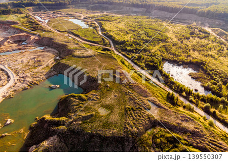 Aerial view of a vast open-pit quarry with turquoise water bodies and rugged terrain. Dirt roads snake through the landscape, bordered by lush green vegetation. Industrial extraction meets natural 139550307