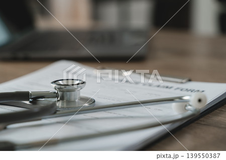 Stethoscope placed on medical claim form on clipboard with blurred laptop background, representing health insurance documentation, medical verification process, patient record review, and healthcare Stethoscope placed on medical claim form on clipboard with blurred laptop background, representing health insurance documentation, medical verification process, patient record review, and healthcare 139550387