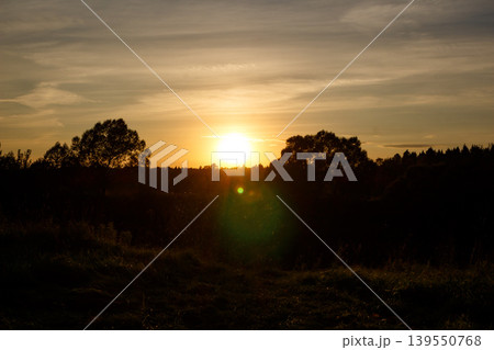 Golden sun dips below the horizon, casting vibrant hues across silhouetted trees and fields. A serene rural twilight scene Golden sun dips below the horizon, casting vibrant hues across silhouetted trees and fields. A serene rural twilight scene 139550768