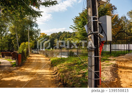 A stout power pole, jampacked with an electrical box and snaking wires. Behind it, a country dirt lane and green trees fade under a clear sky 139550815