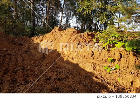 Earthen trench with a freshly dug mound of soil, illuminated by sunlight. Trees in the background offer a natural backdrop, highlighting construction or landscaping Earthen trench with a freshly dug mound of soil, illuminated by sunlight. Trees in the background offer a natural backdrop, highlighting construction or landscaping 139550834