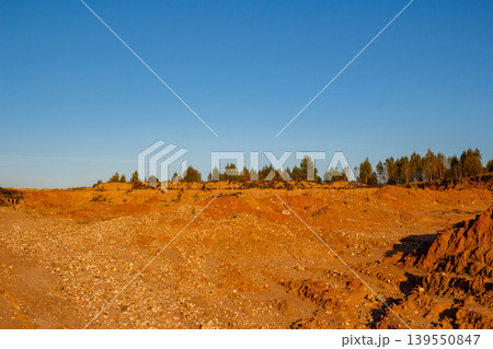 Vast open-pit quarry, reddish-orange sand and gravel terrain under a clear blue sky. Distant green trees line the horizon, showing an industrial landscape Vast open-pit quarry, reddish-orange sand and gravel terrain under a clear blue sky. Distant green trees line the horizon, showing an industrial landscape 139550847