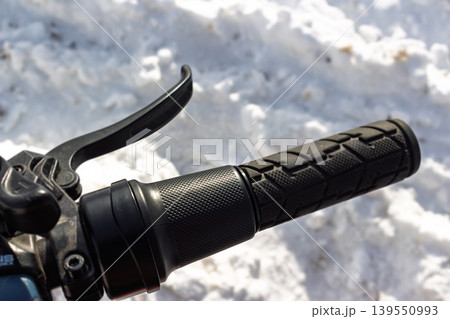 Electric bike's right handlebar shows a rugged grip and half-twist throttle. Blurred white snow forms a chilly backdrop, perfect for winter e-cycling adventures 139550993