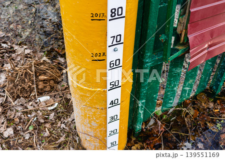 Yellow metal pole featuring a white water level gauge with historical flood marks from 2013 and 2023, situated at a rural entrance during the spring thaw season in Russia 139551169